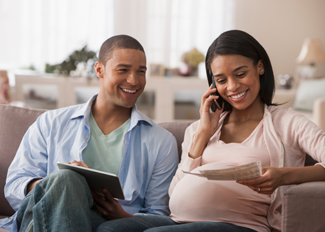 Happy black couple sitting on the couch. Woman is pregnant and talking on the phone. Man sits next to her with a pad and pen taking notes.
