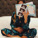 Woman sitting on bed holding pregnant belly and reading with face hidden behind pregnancy books