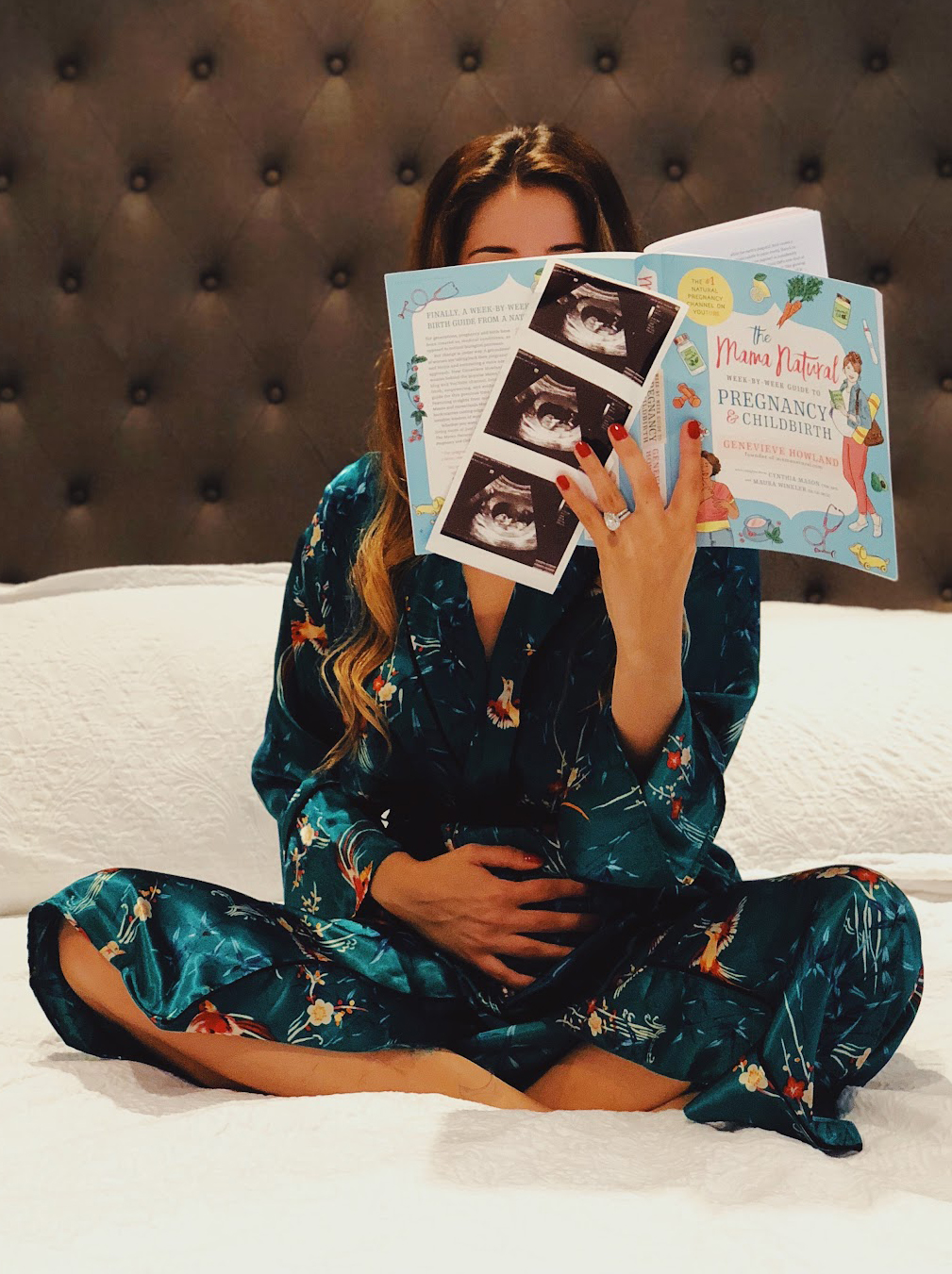 Woman sitting on bed holding pregnant belly and reading with face hidden behind pregnancy books